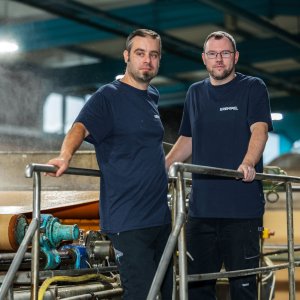 Two men standing in a production area on a platform with steam in the background looking into the camera